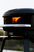 Gozney pizza oven with visible flames inside, set against a blurred outdoor background.