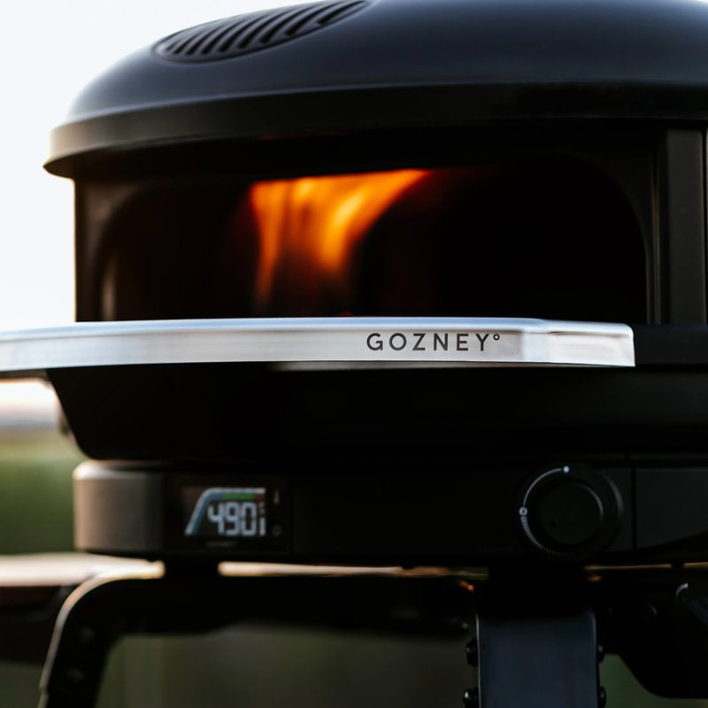 Gozney pizza oven with visible flames inside, set against a blurred outdoor background.