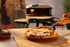 Person cutting a pizza in front of a portable pizza oven with people in the background.
