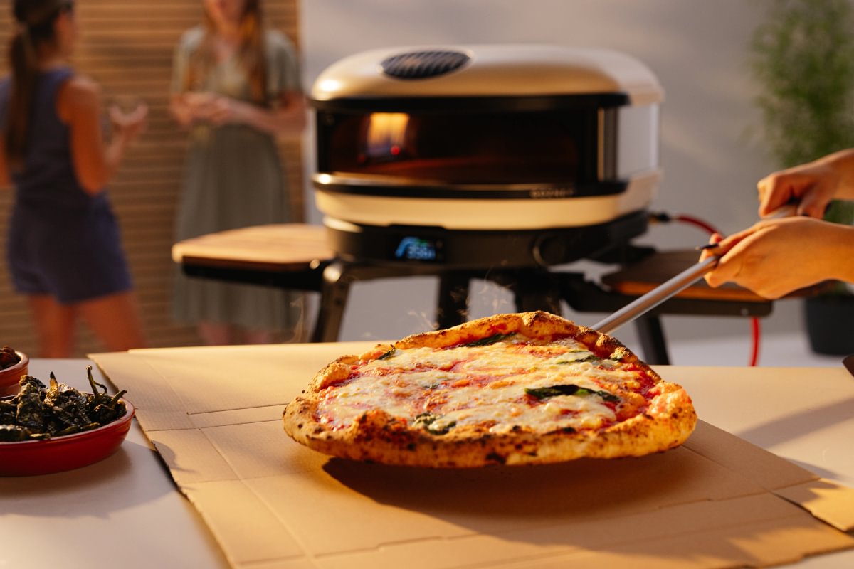 Person cutting a pizza in front of a portable pizza oven with people in the background.