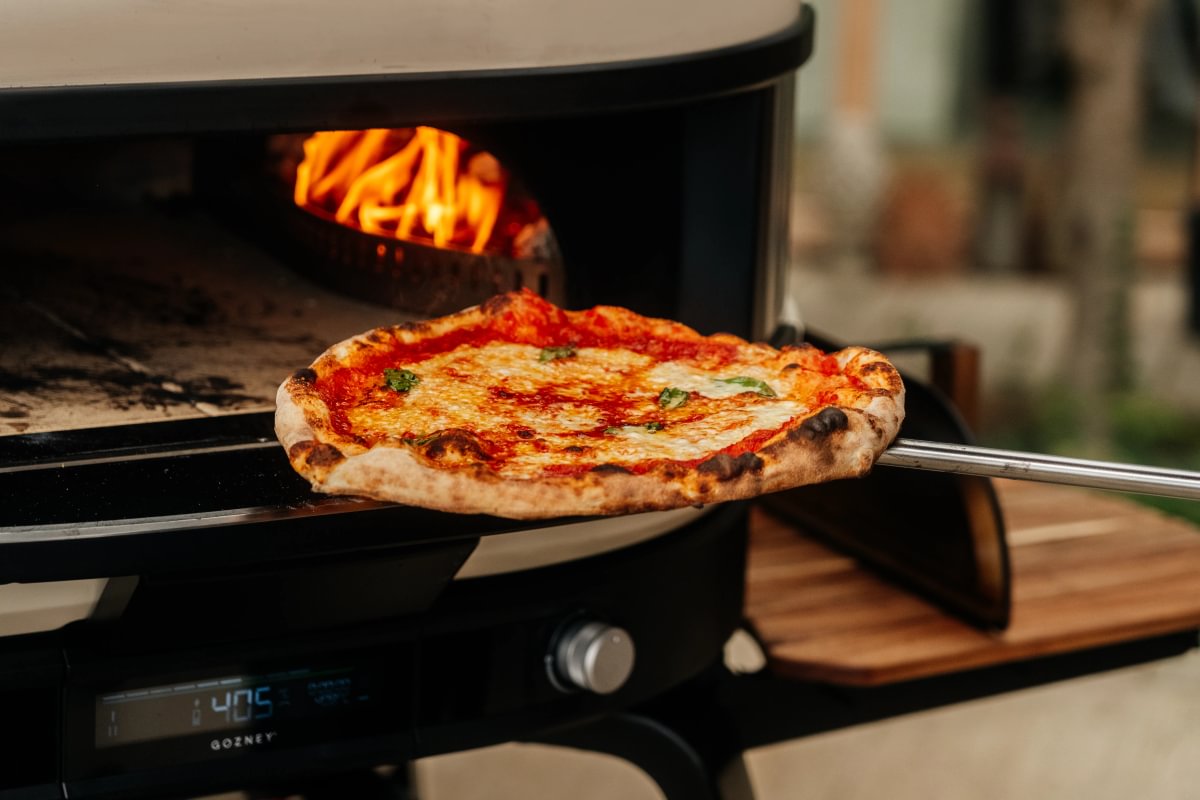 Wood-fired pizza being taken out of a portable pizza oven with flames visible.