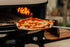 Wood-fired pizza being taken out of a portable pizza oven with flames visible.