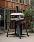 Portable outdoor pizza oven on a stand in front of a wooden building.
