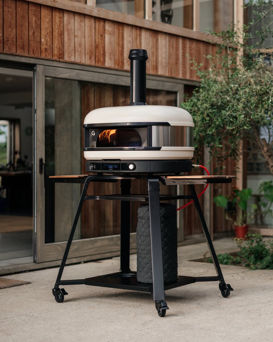 Portable outdoor pizza oven on a stand in front of a wooden building.