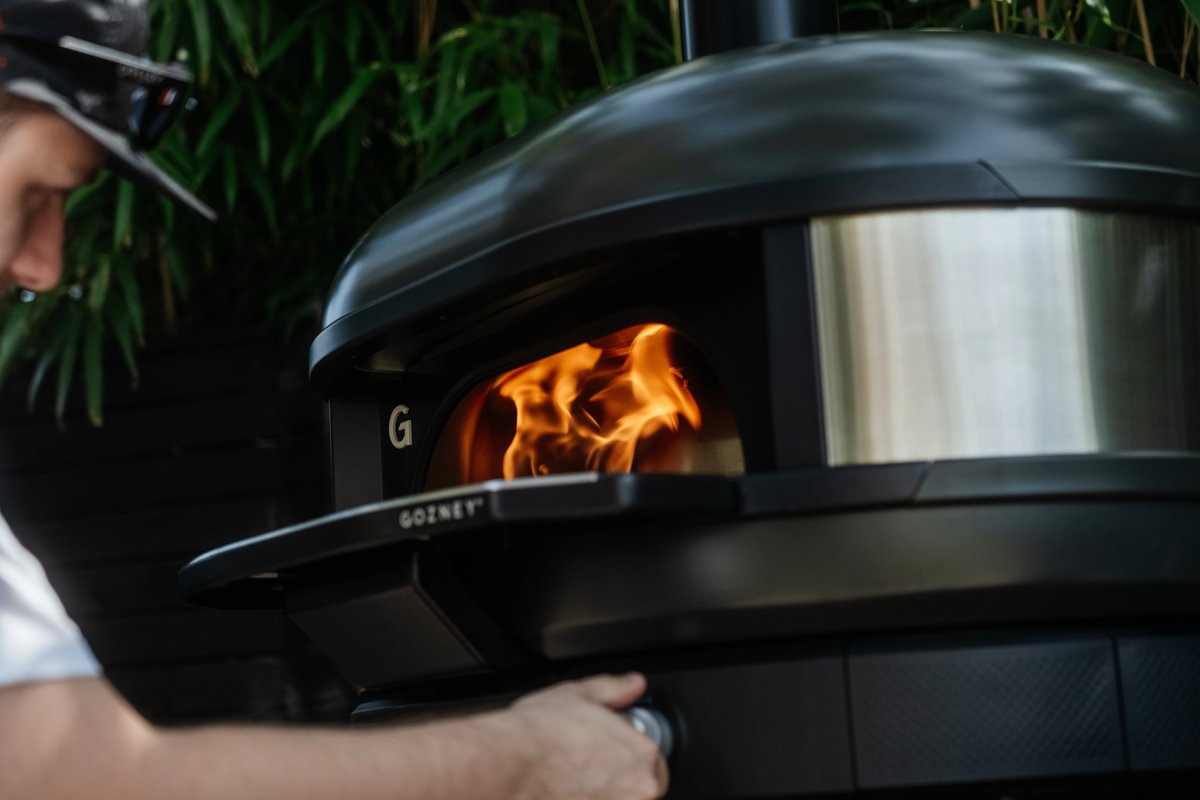 Person interacting with a black outdoor pizza oven with visible flames.