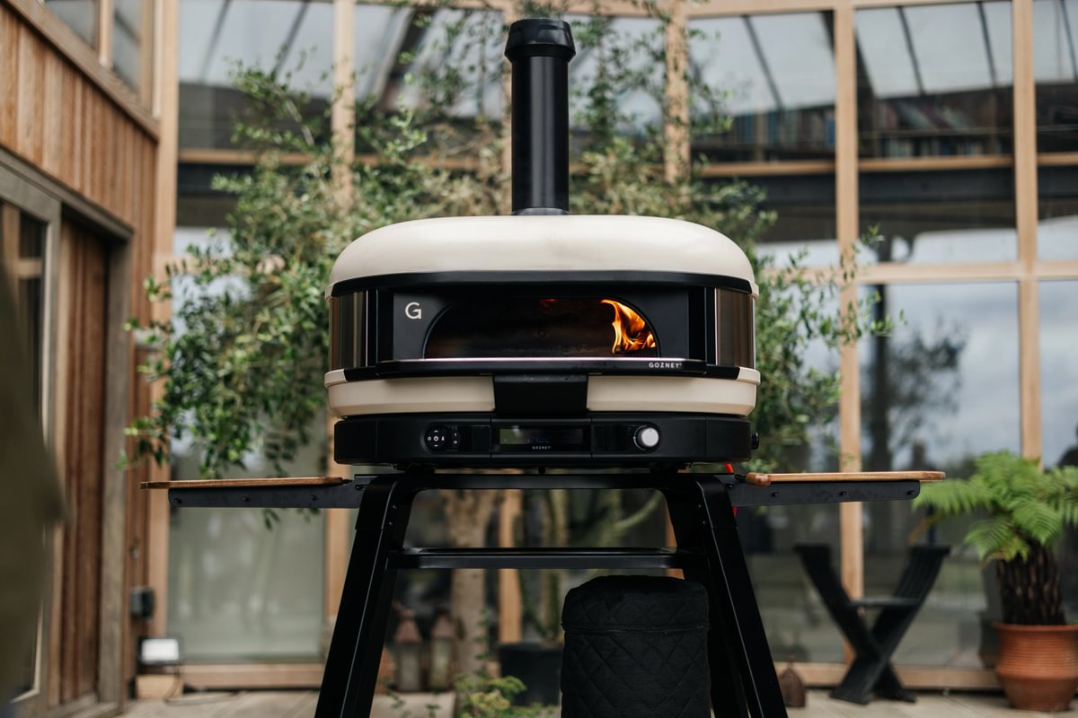 Outdoor pizza oven with visible flame inside, set against a wooden and glass background.