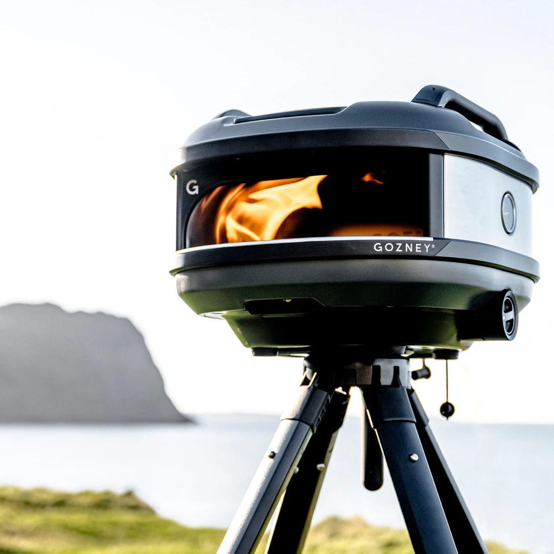 Gozney stove on a tripod with flames inside, set against a natural landscape.